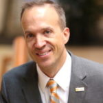 Photo of a male with pale skin wearing a grey suit and an orange and white stripped tie.